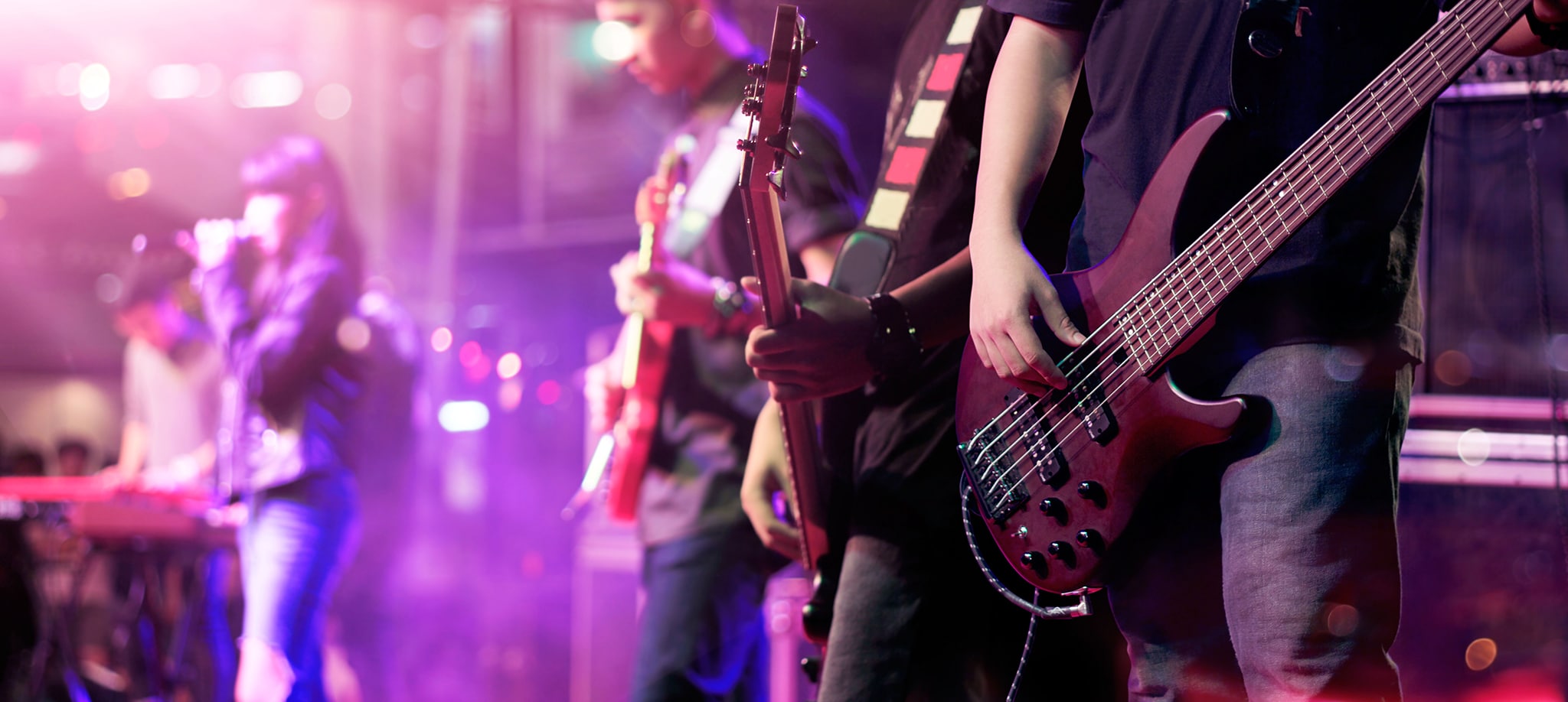 Musiker auf der Bühne. Nahaufnahme eines Bassisten, der im Vordergrund Gitarre spielt, während andere Bandmitglieder im Hintergrund E-Gitarre und Keyboard spielen. Die Szene wird von bunten Bühnenlichtern beleuchtet.