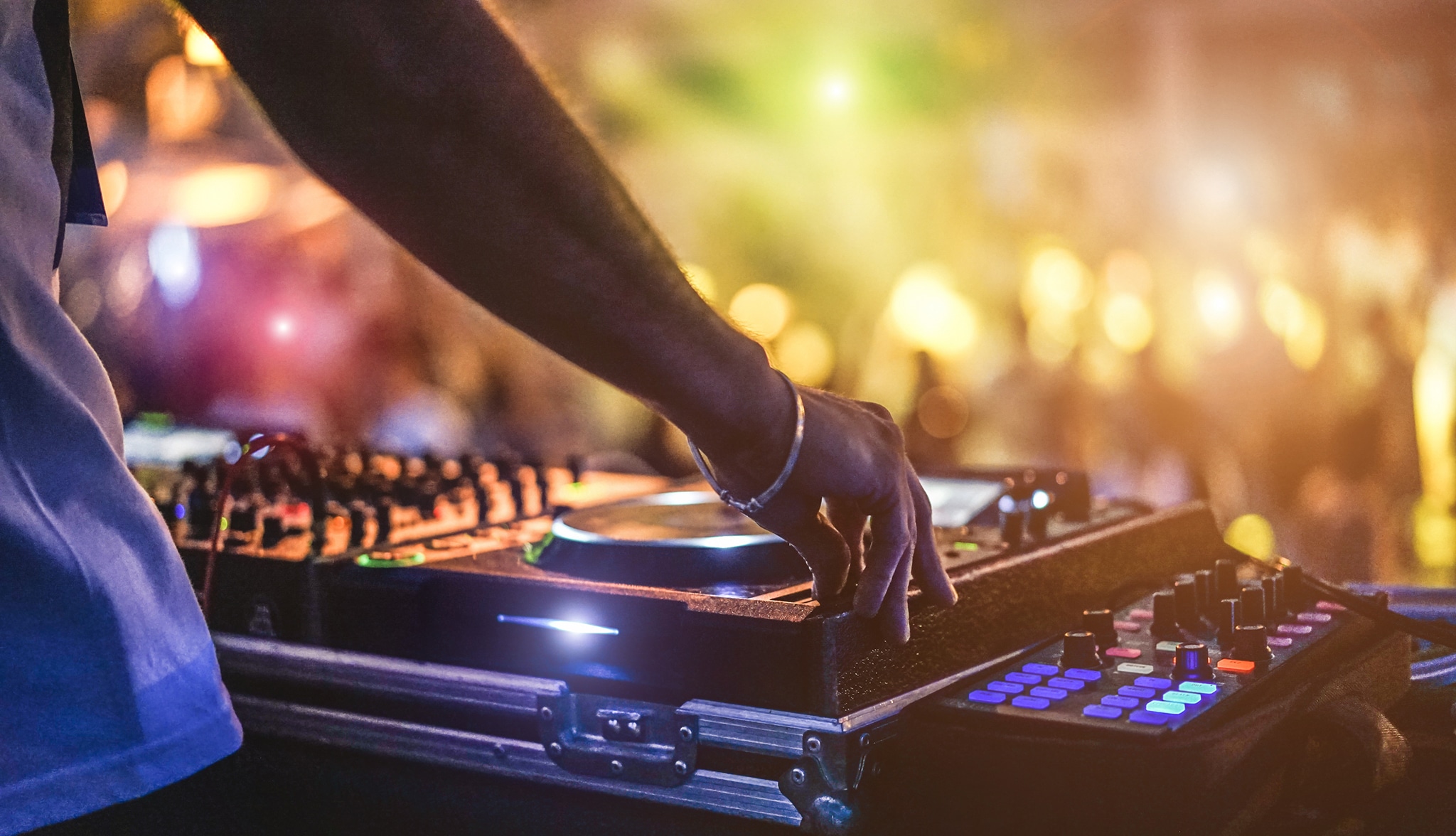 Ein DJ mixt Musik bei einem Auftritt. Die Szene ist schwach beleuchtet, im Hintergrund sind bunte Lichter zu sehen. Die Hand des DJs liegt auf dem Plattenteller, umgeben von verschiedenen Audiogeräten und leuchtenden Knöpfen.