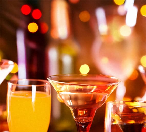 Nahaufnahme verschiedener Gläser mit verschiedenfarbigen Getränken, darunter ein Martini-Glas im Vordergrund. Im Hintergrund sind verschwommene, leuchtend orange und gelbe Bokeh-Lichter zu sehen.