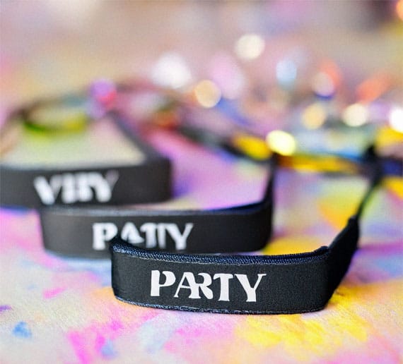 Nahaufnahme von drei schwarzen Armbändern auf buntem Hintergrund. Der sichtbare Text auf den Armbändern lautet „VERY“, „PARTY“ und auf dem letzten ist ein teilweise verdecktes Wort zu sehen. Der Hintergrund ist von verschwommenen Lichtern und einem Mix aus Pastellfarben geprägt.